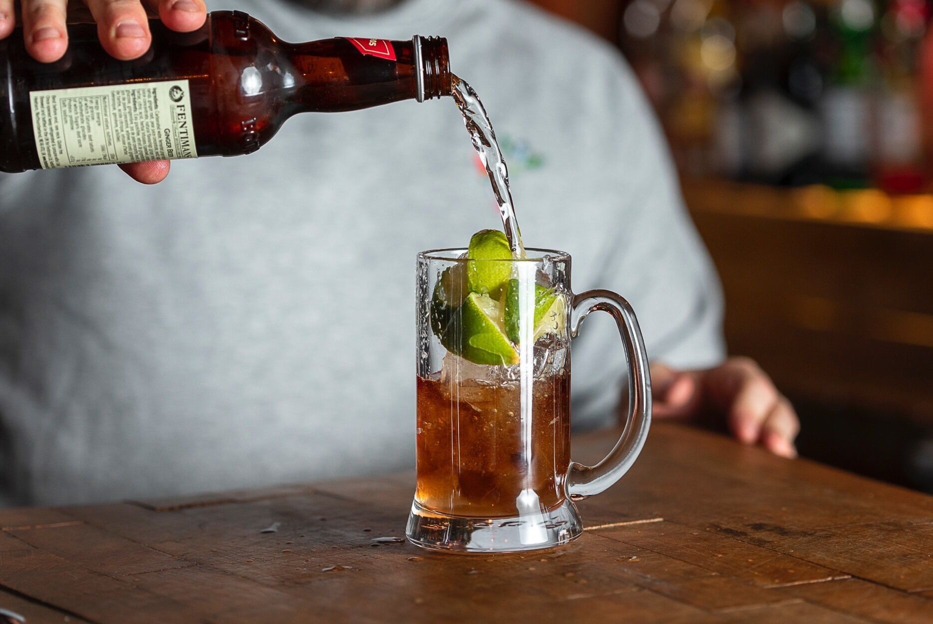 Una persona vertiendo un líquido oscuro de una botella en un vaso de vidrio transparente con asa, con hielo y rodajas de limón.