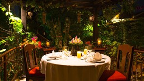 Un área de comedor al aire libre con una mesa redonda preparada para dos personas, rodeada de abundante vegetación y faroles decorativos.