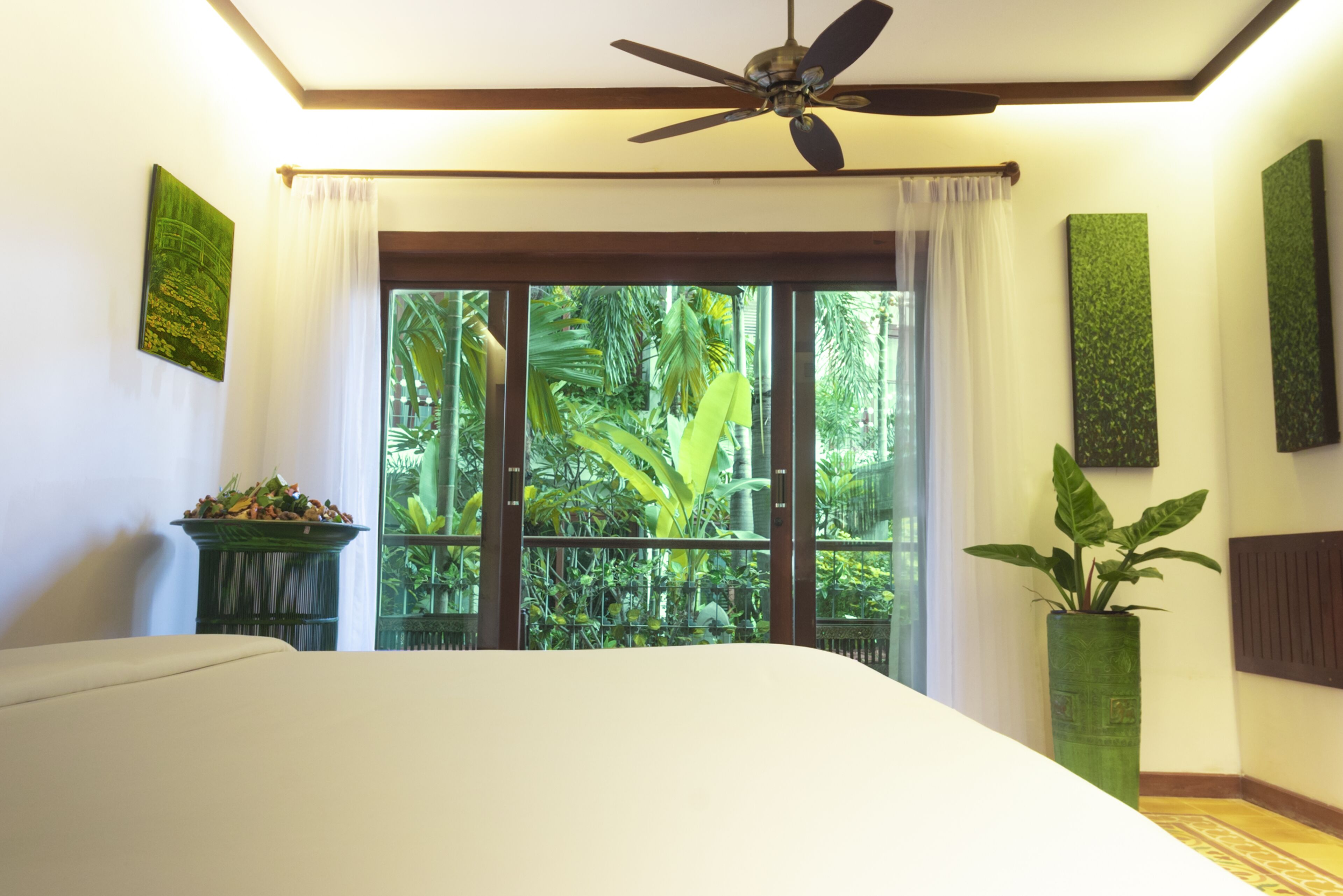 A bedroom with a bed, a ceiling fan, a window with curtains, a potted plant, and a wall-mounted artwork.