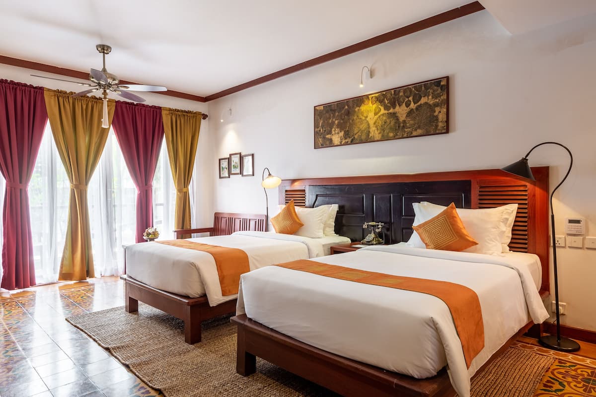 Ein Hotelzimmer mit zwei Betten, einem Deckenventilator, einem großen Gemälde an der Wand und einer Stehlampe.