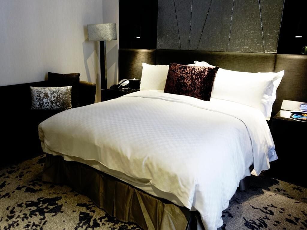 A neatly made bed with a white comforter and two pillows, a bedside lamp, and a nightstand with a phone.