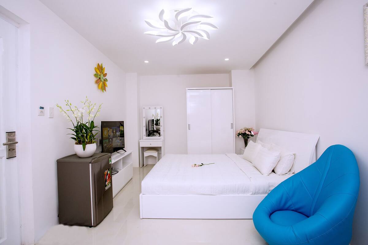 Une chambre moderne avec un lit blanc, une chaise bleue, une commode, une coiffeuse avec un miroir et un luminaire au plafond.