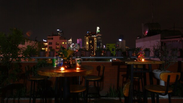 A rooftop terrace with tables and chairs, potted plants, and a cityscape view at night.