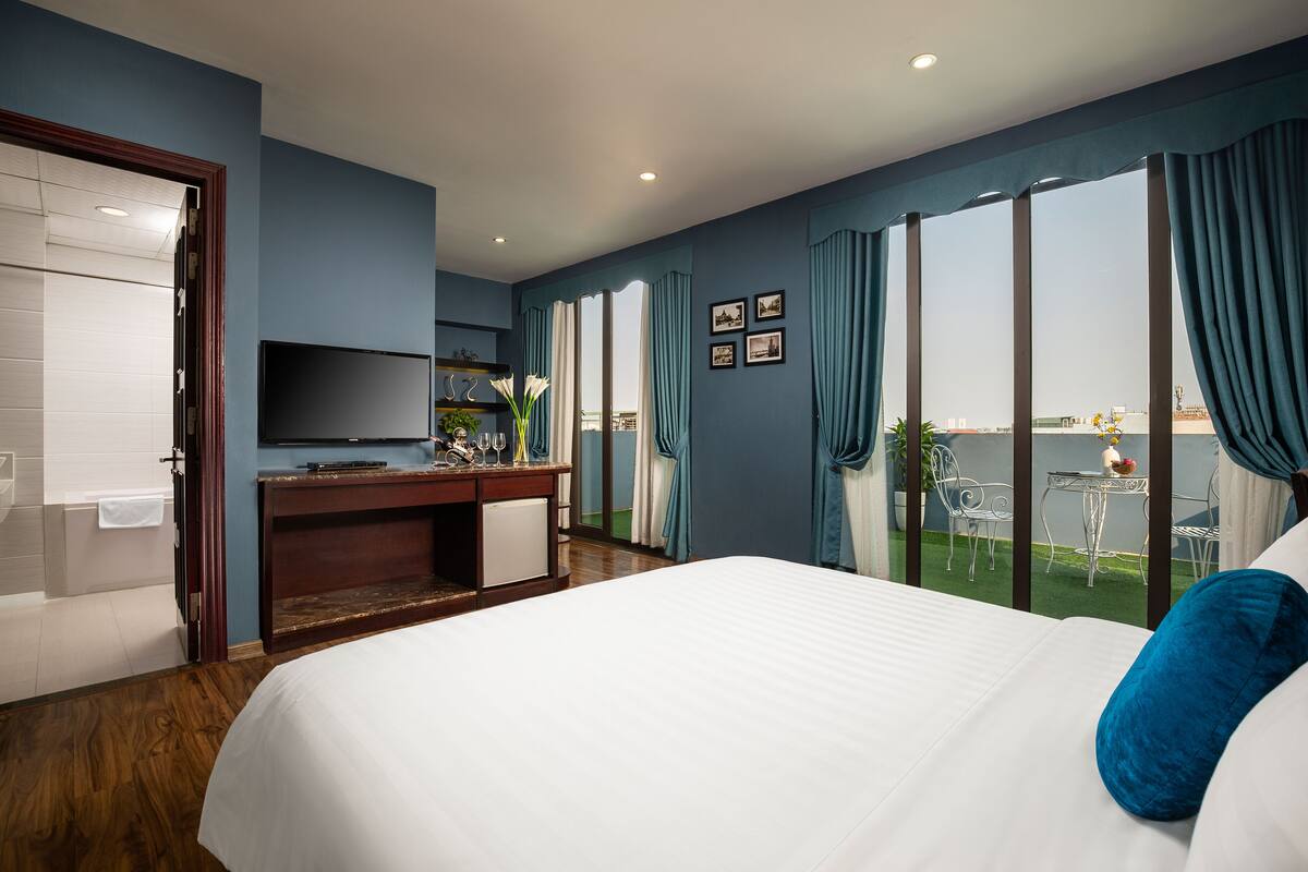 Une chambre d’hôtel avec un grand lit, un bureau avec une télévision et une vue sur un balcon aménagé avec des sièges d’extérieur.