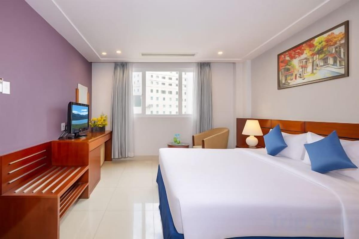 A hotel room with a bed, a desk with a computer, a chair, a lamp, and a window with curtains.