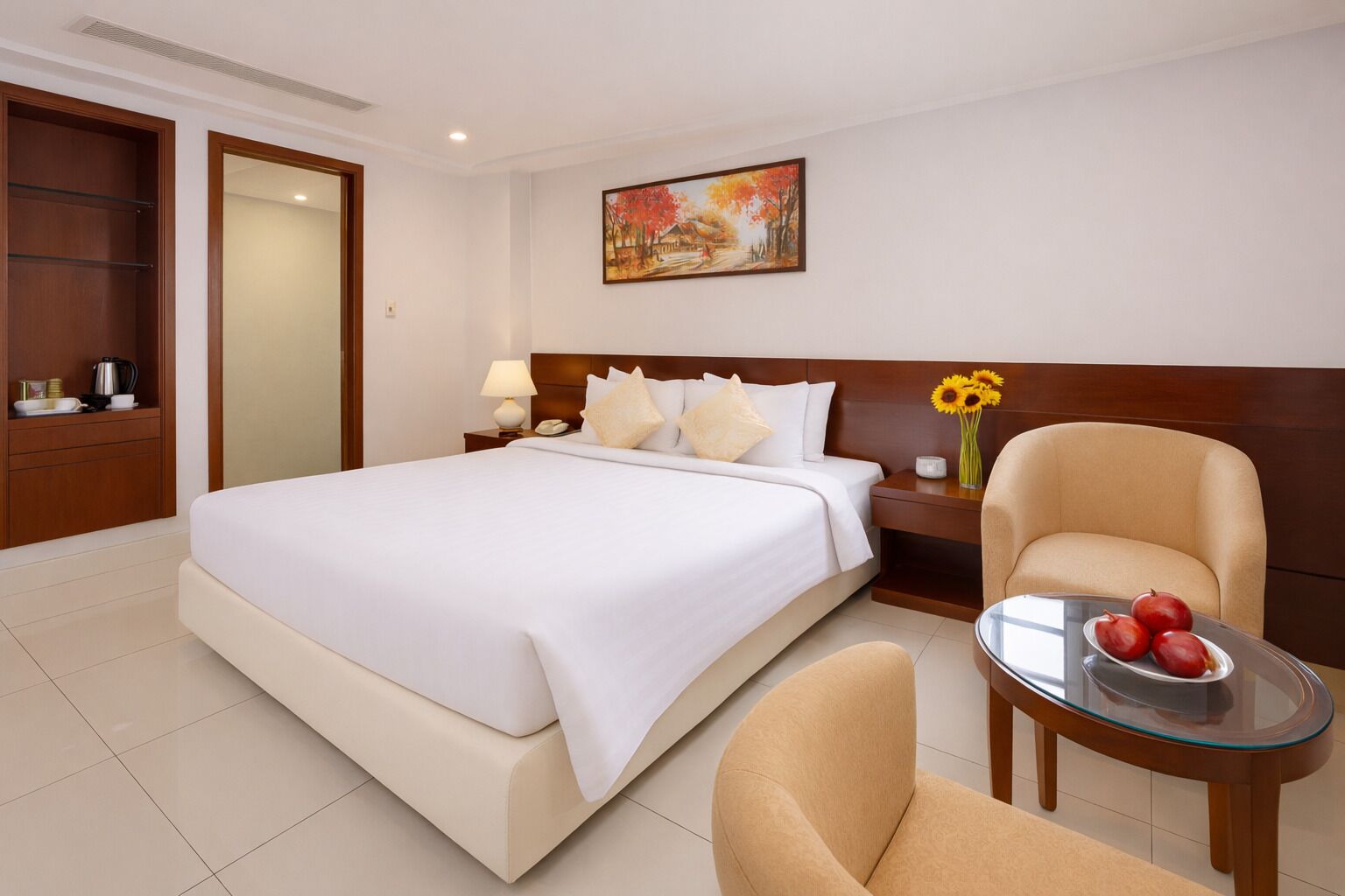 Une chambre d’hôtel avec un grand lit, une table de chevet avec une lampe, une chaise, une petite table avec des fruits et un tableau au mur.