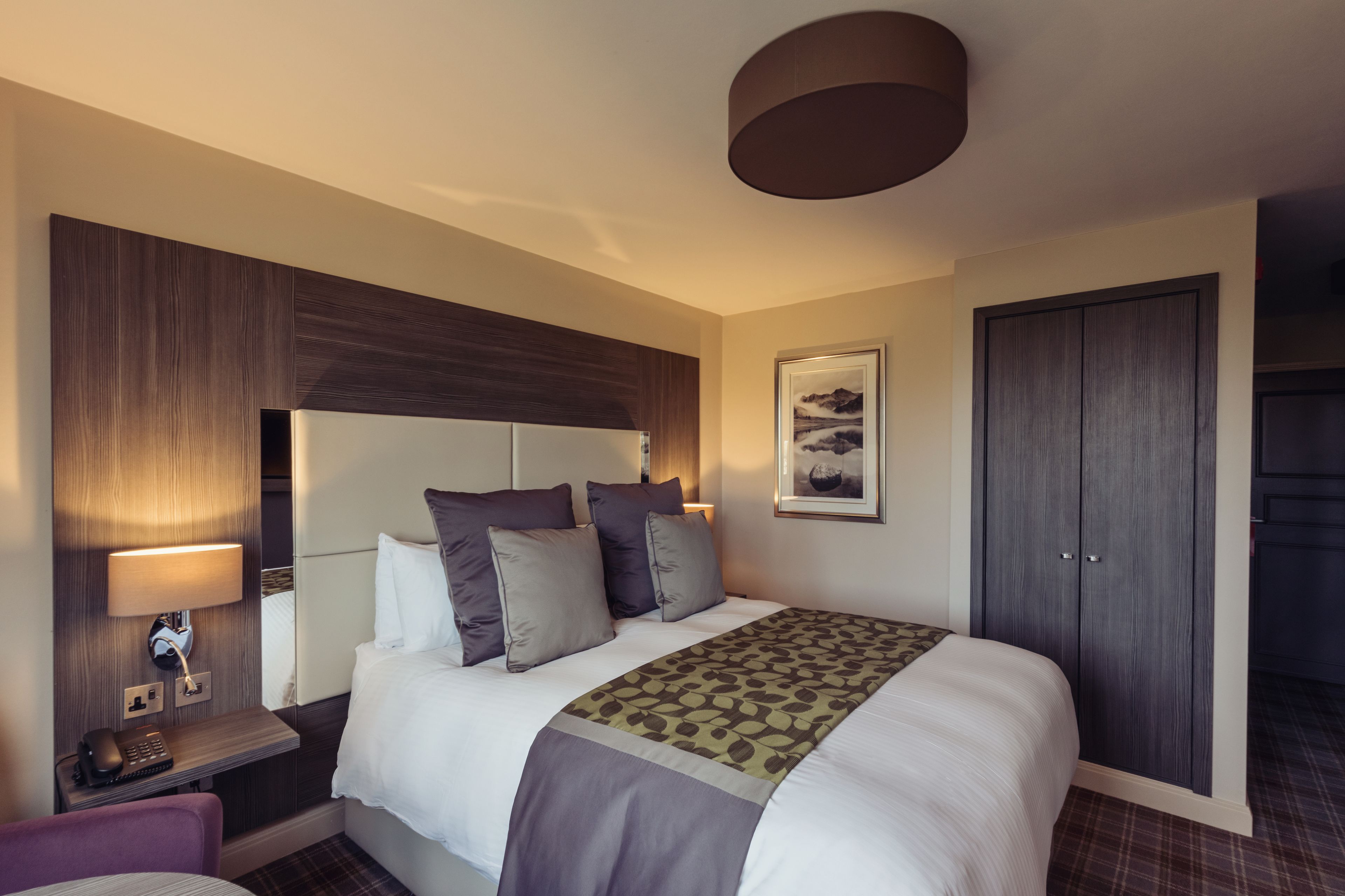 A hotel room with a large bed, a bedside table with a lamp, a chair, and a wardrobe.