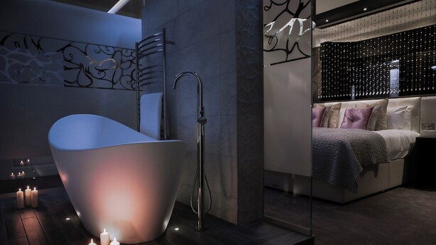 A modern bathroom with a freestanding bathtub, a glass-enclosed shower area, and a bed with a view of the cityscape. - Abbey House Hotel (Barrow-in-Furness)