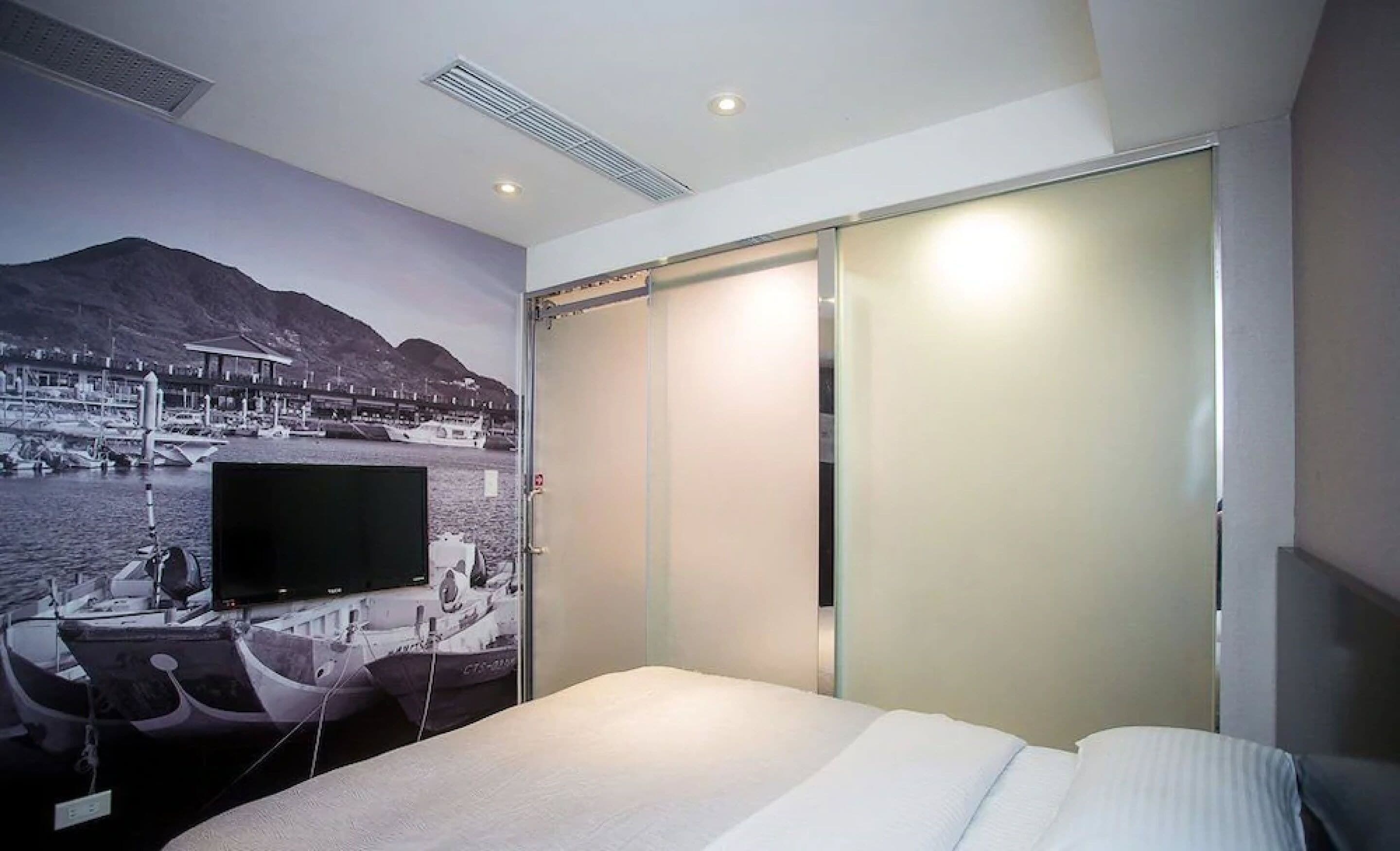 Quarto de hotel com cama, televisão na parede, porta deslizante e um mural de um porto de recreio.