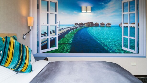 A bedroom with a large window revealing a beach scene with overwater bungalows. - E House Hotel (Taipei)