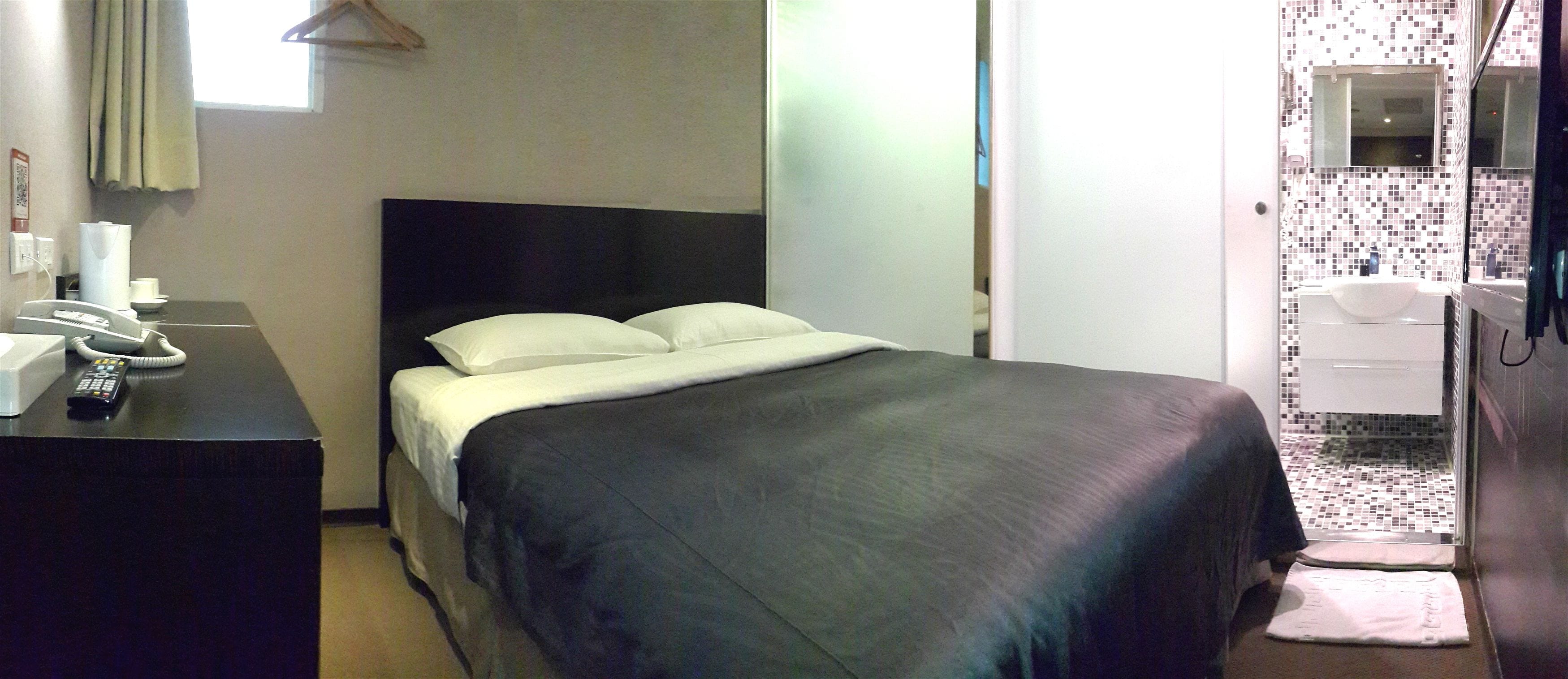 Hotelkamer met een bed, een bureau met telefoon en afstandsbediening, een badkamer met wastafel en spiegel, en een raam met gordijnen.
