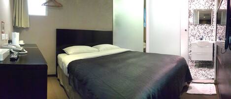 Hotelkamer met een bed, een bureau met telefoon en afstandsbediening, een badkamer met wastafel en spiegel, en een raam met gordijnen.