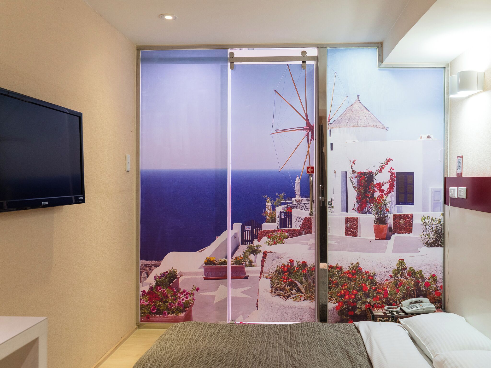A bedroom with a large window offering a view of a coastal scene, a bed, a bedside table, and a flat-screen TV mounted on the wall.