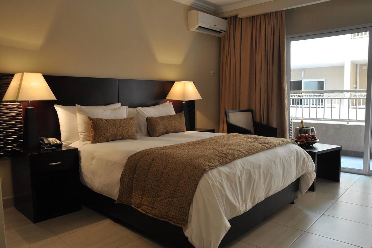 Ein Hotelzimmer mit einem großen Bett, zwei Nachttischen mit Lampen, einem kleinen Tisch mit Obst und Blick auf den Balkon.