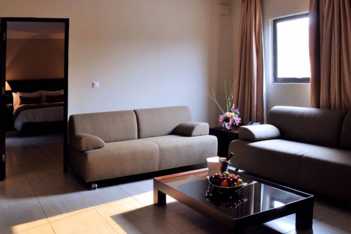 Ein modernes Hotelzimmer mit einem beigen Sofa, einem Couchtisch mit Früchten, einer Blumenkomposition und einem Bett im Hintergrund.
