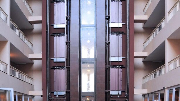 A modern hotel lobby with multiple dining tables and chairs, large windows, and a central elevator. - Hotel Royal (Kinshasa)