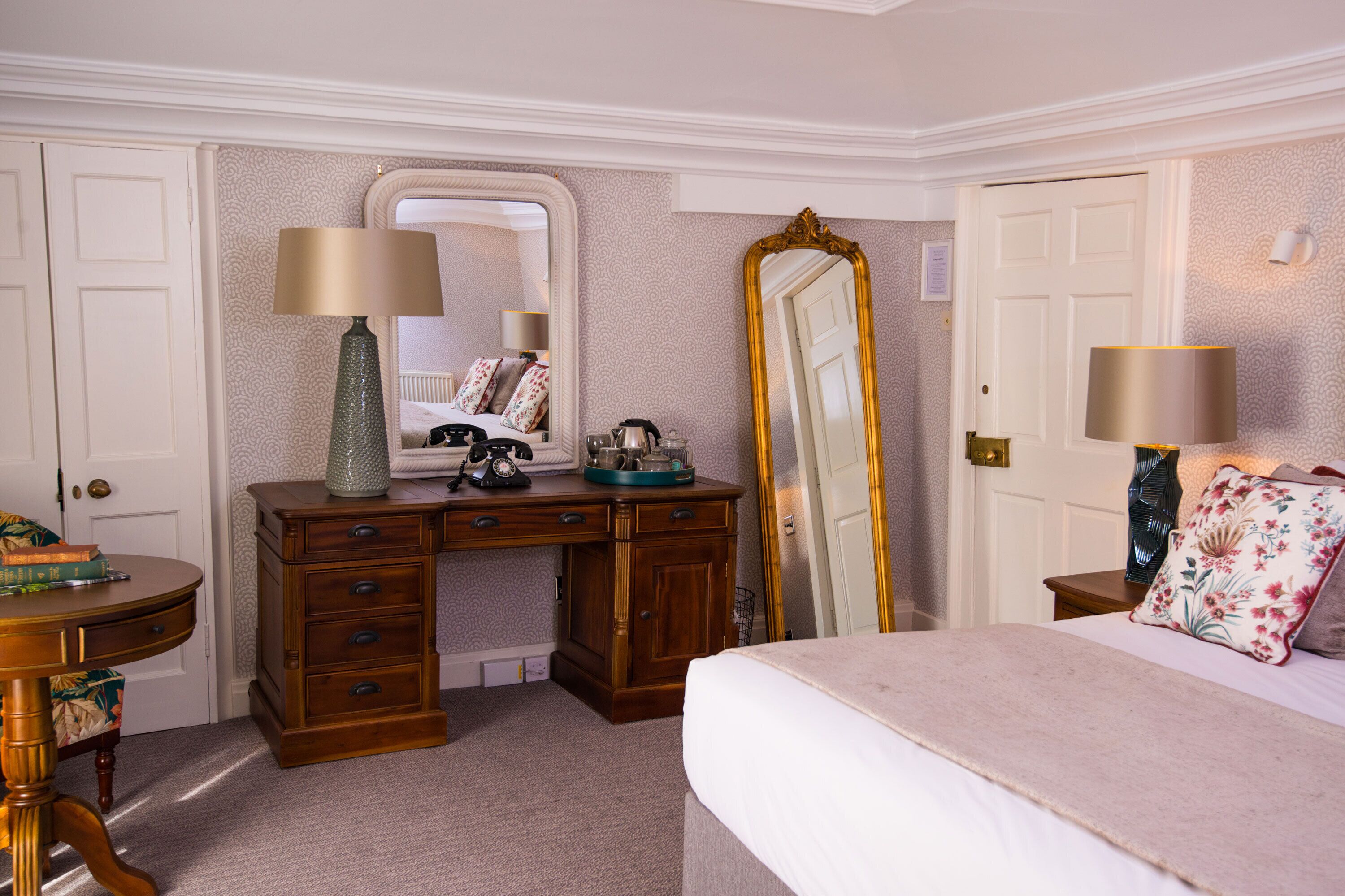 A bedroom with a large bed, a dresser with a mirror, a desk with a lamp, and a door.