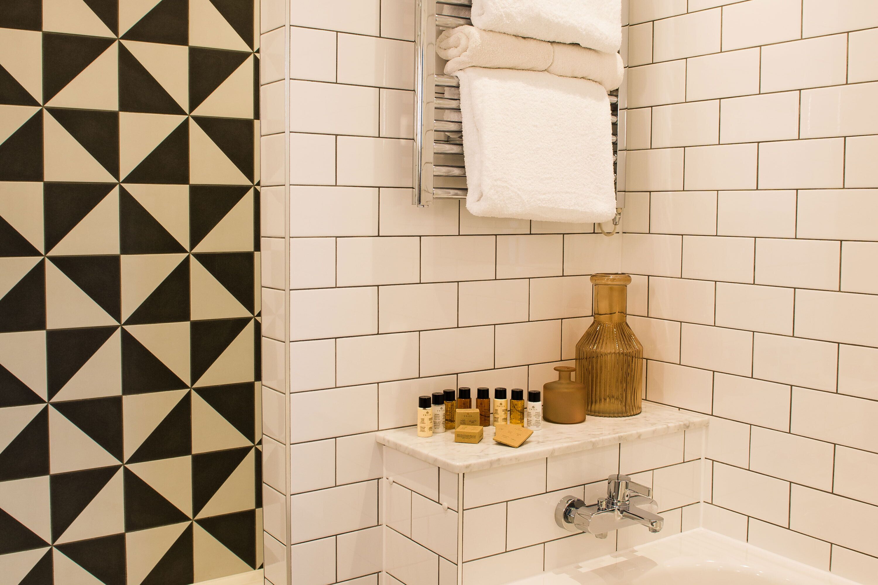 Casa de banho com azulejos brancos, padrão geométrico preto e branco na parede, toalheiro, prateleira com artigos de higiene pessoal e uma banheira.