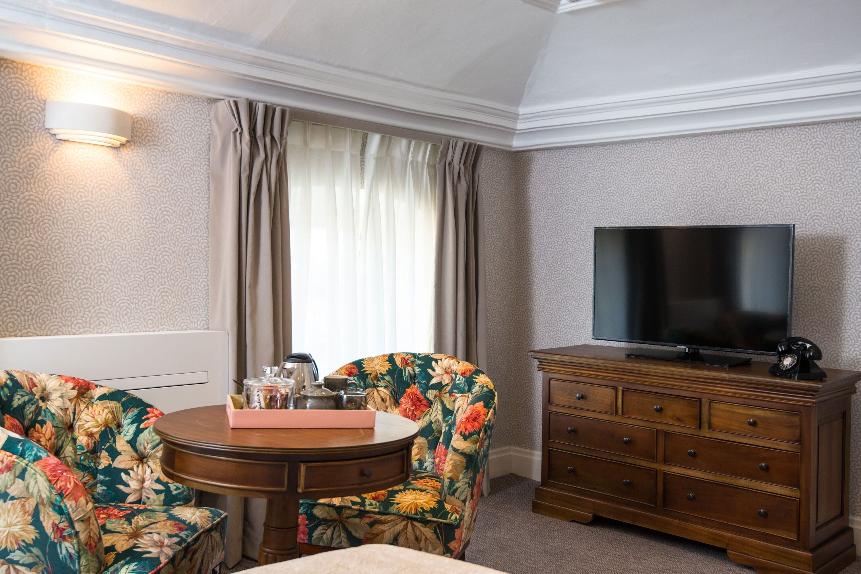 A hotel room with a round wooden table, floral armchairs, a TV, and a wooden dresser.