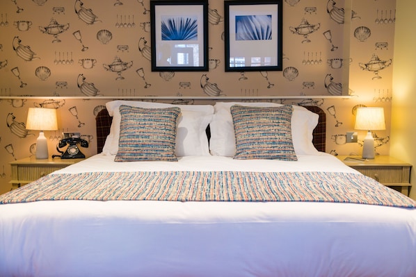 Cama bem arrumada com almofadas estampadas, um telefone antigo na mesa de cabeceira e quadros nas paredes.