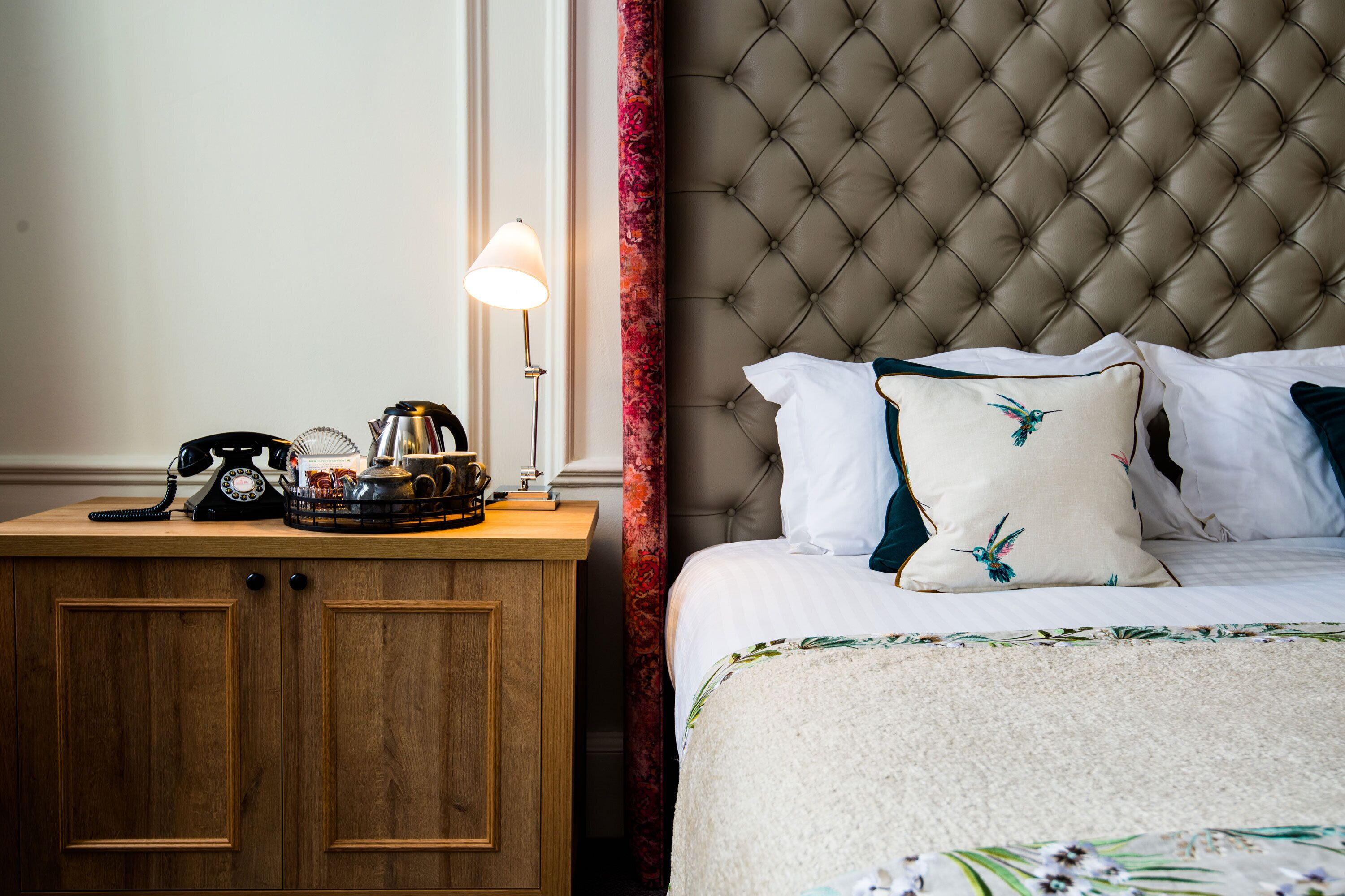 Una habitación de hotel con una mesita de noche de madera, un teléfono, una lámpara y una cama con cabecera capitonada y ropa de cama con motivos florales.