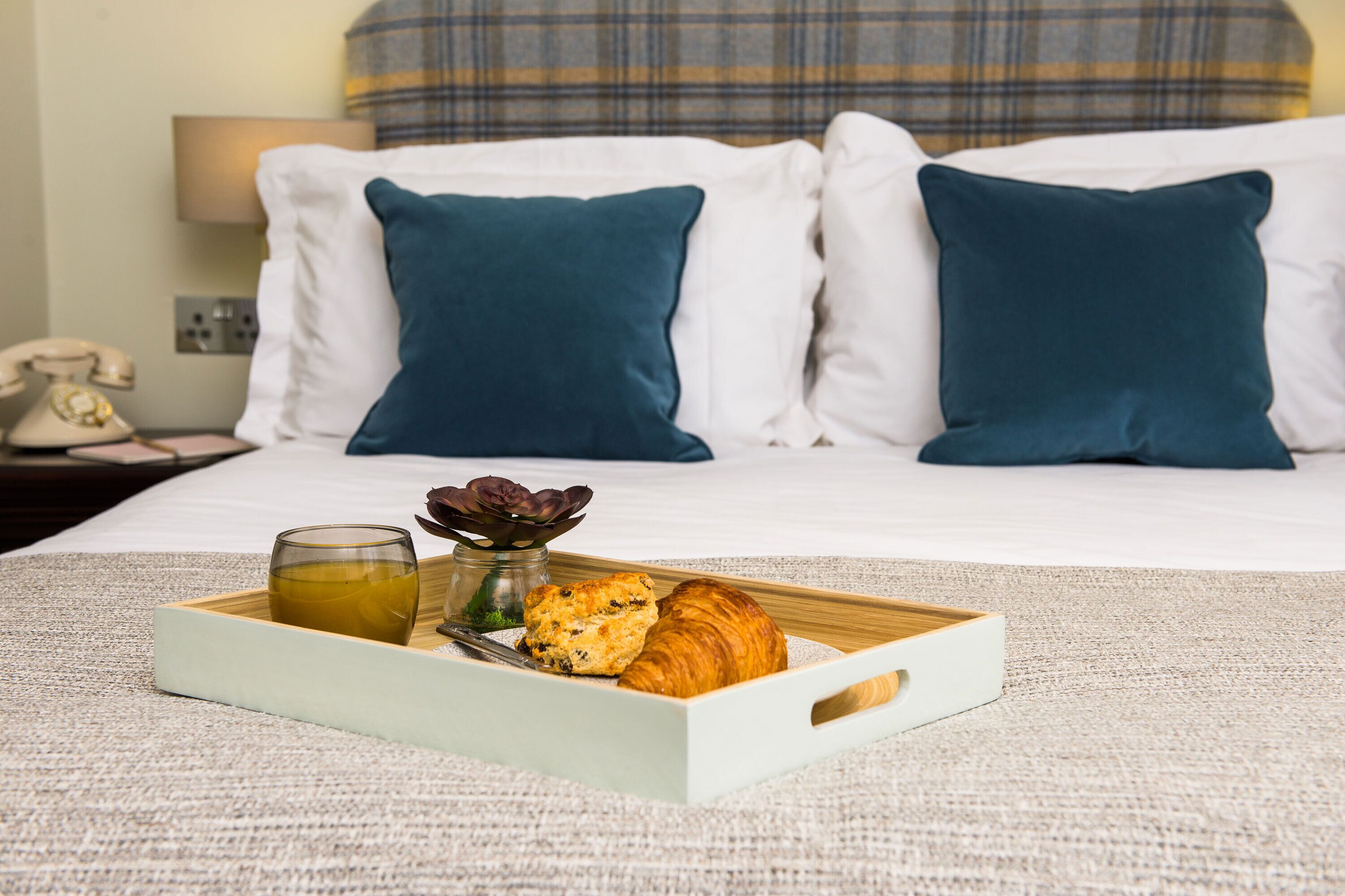 A neatly made breakfast tray with croissants and a glass of juice on a bed with white and dark blue pillows, a lamp, and a telephone on the nightstand.