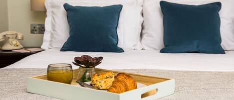 A neatly made breakfast tray with croissants and a glass of juice on a bed with white and dark blue pillows, a lamp, and a telephone on the nightstand.