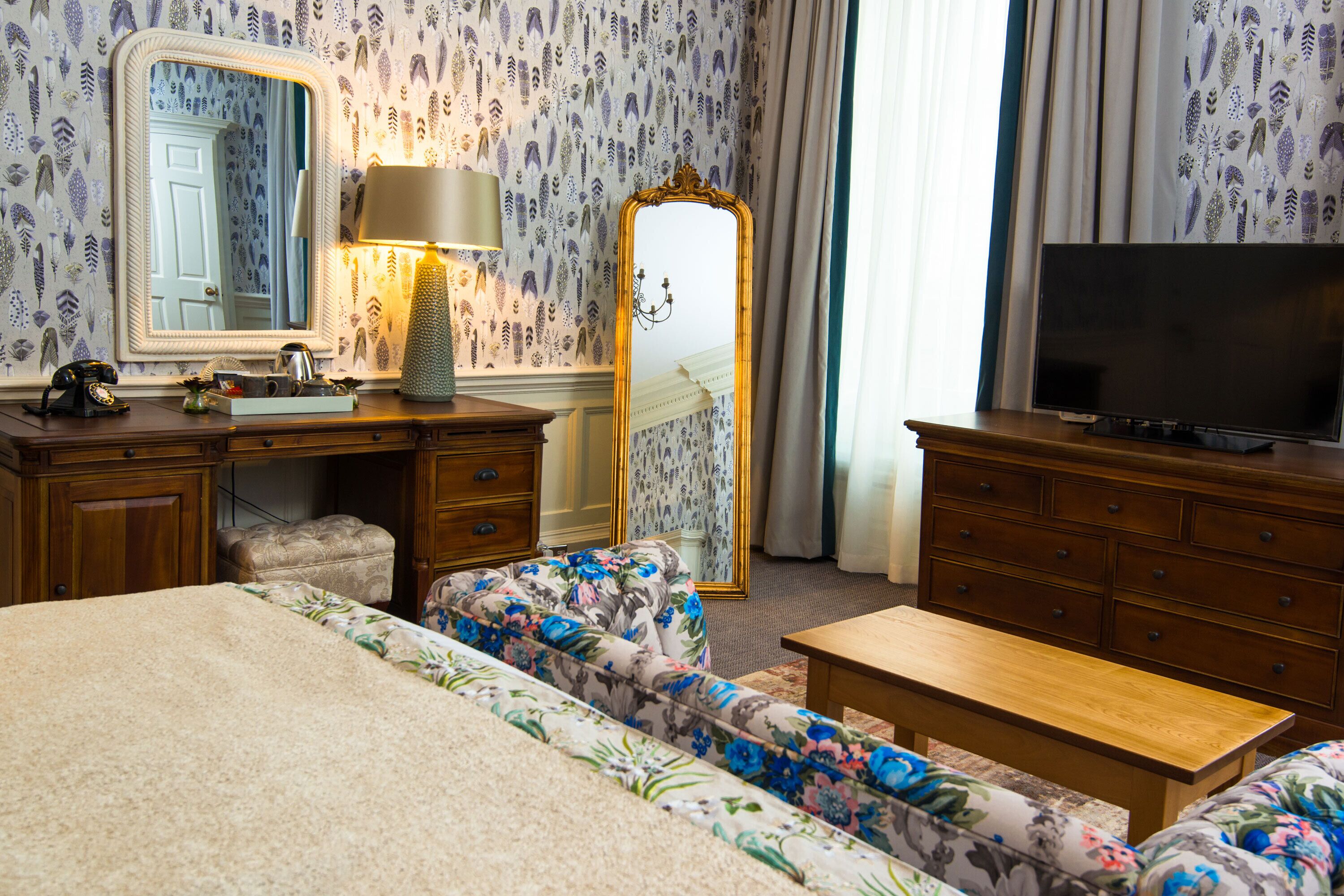 A hotel room with a bed, a sofa, a TV, a dresser, a mirror, a lamp, and a window with curtains.