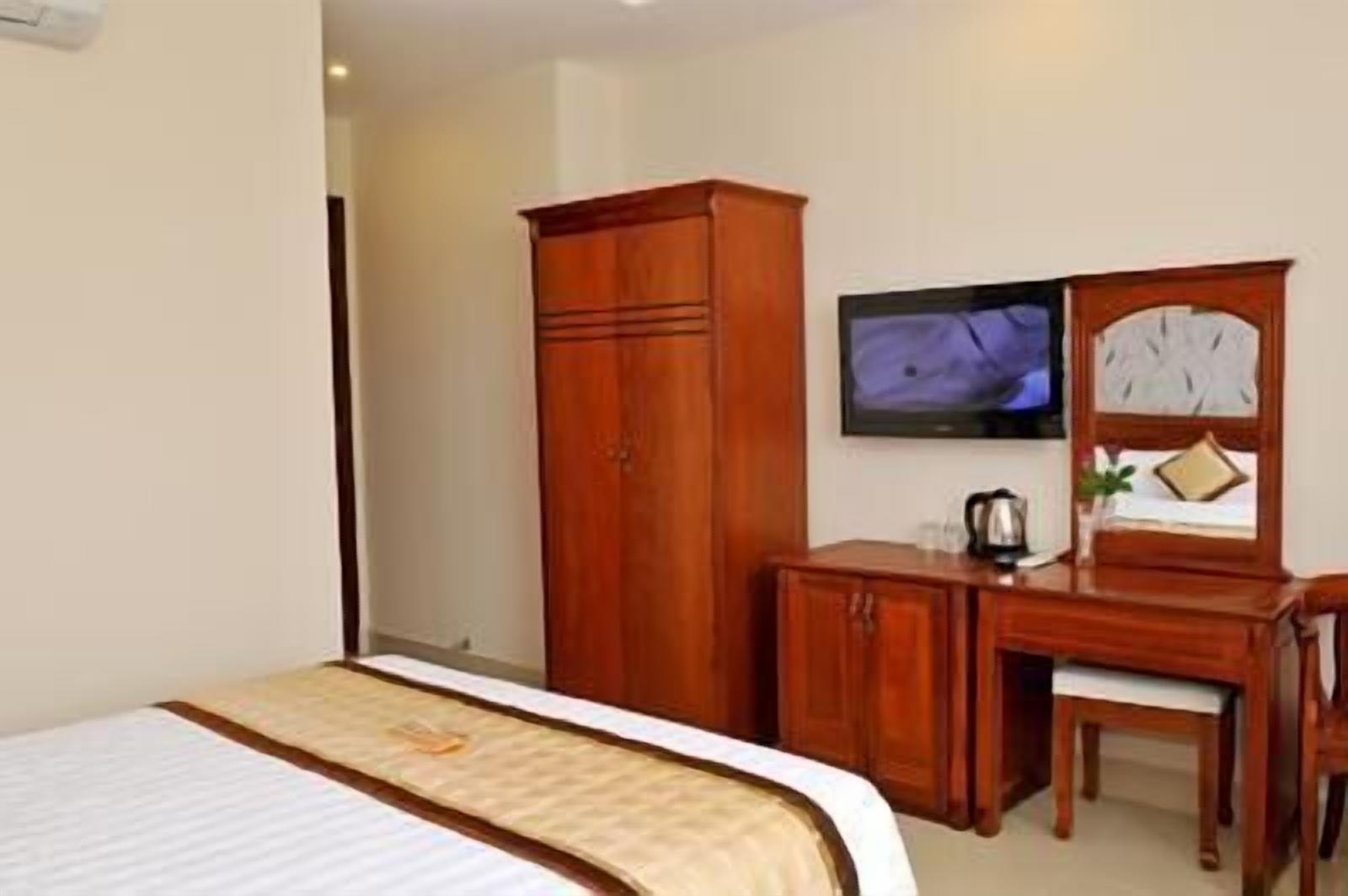 Een hotelkamer met een bed, een houten kledingkast, bureau, stoel en een televisie aan de muur.
