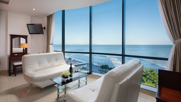 Quarto de hotel moderno com uma grande janela panorâmica, um sofá de couro branco, uma mesa de centro de vidro e uma televisão de tela plana presa à parede.