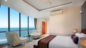 Quarto de hotel moderno com uma cama grande, uma poltrona branca, uma mesa de vidro e vista para o oceano.