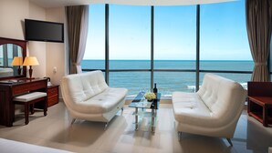 Quarto de hotel com uma grande janela com vista para o oceano, dois sofás de couro branco, uma pequena mesa com um vaso de flores, uma escrivaninha com uma luminária e uma televisão de tela plana na parede.