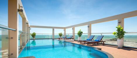 Piscina infinita en la azotea con agua azul, rodeada de una terraza de madera y sillones de descanso, con vistas al océano.