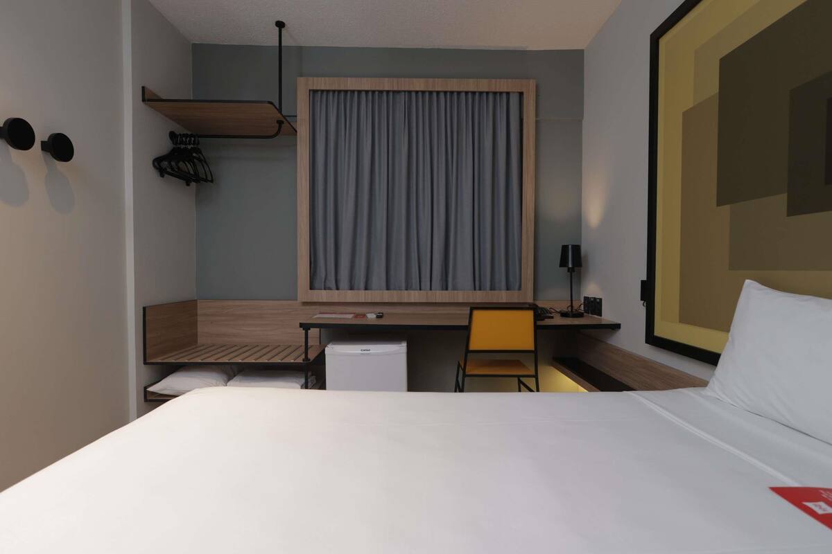 Habitación de hotel con cama, escritorio, silla, refrigerador pequeño, lámpara y ventana grande con cortinas.