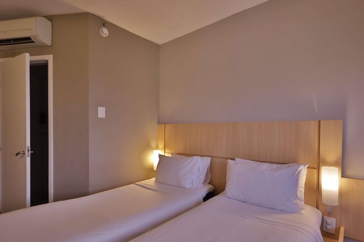 Habitación de hotel con dos camas, cabecero de madera, lámparas de noche y un equipo de aire acondicionado.