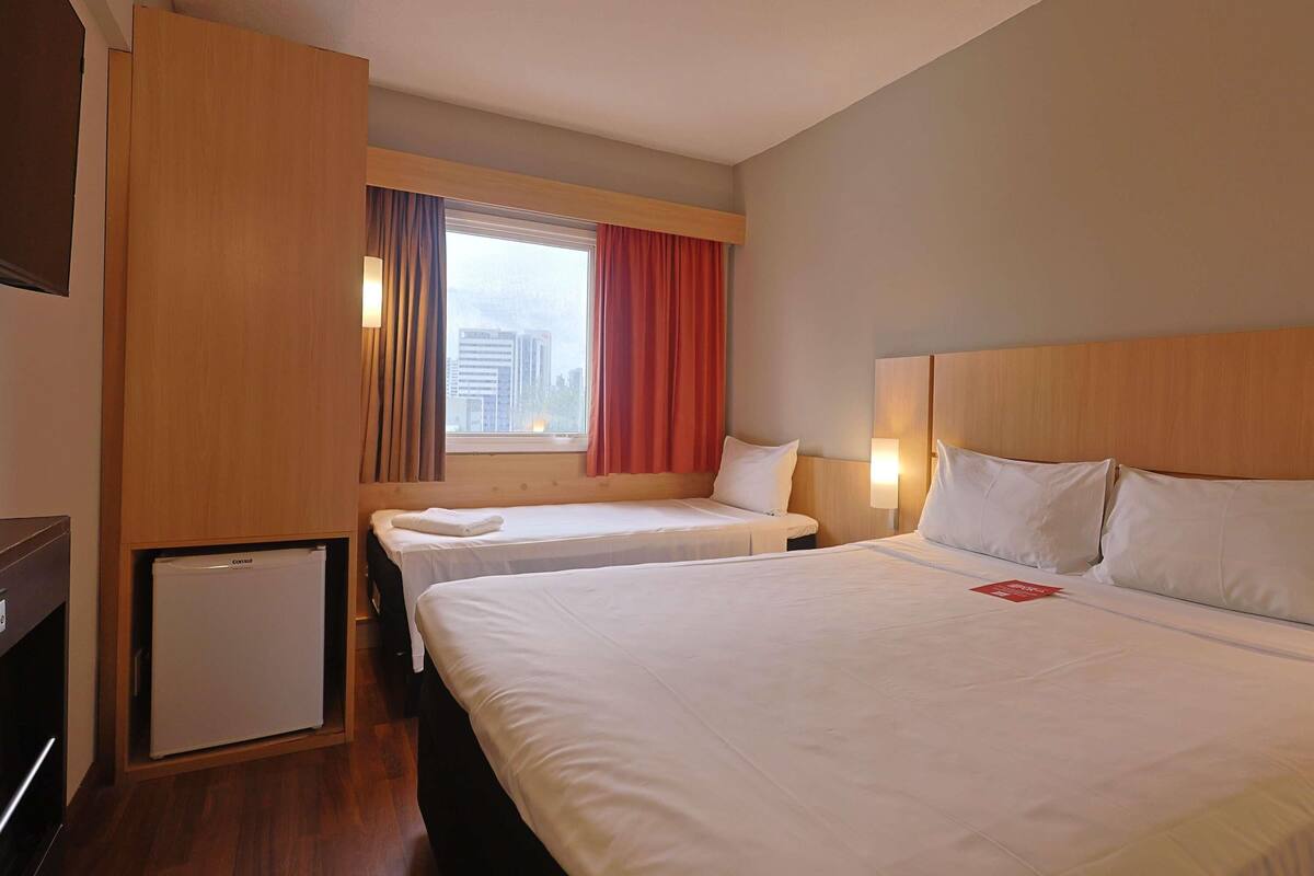 Habitación de hotel con dos camas, un refrigerador pequeño, una ventana con cortinas y una lámpara fijada a la pared.