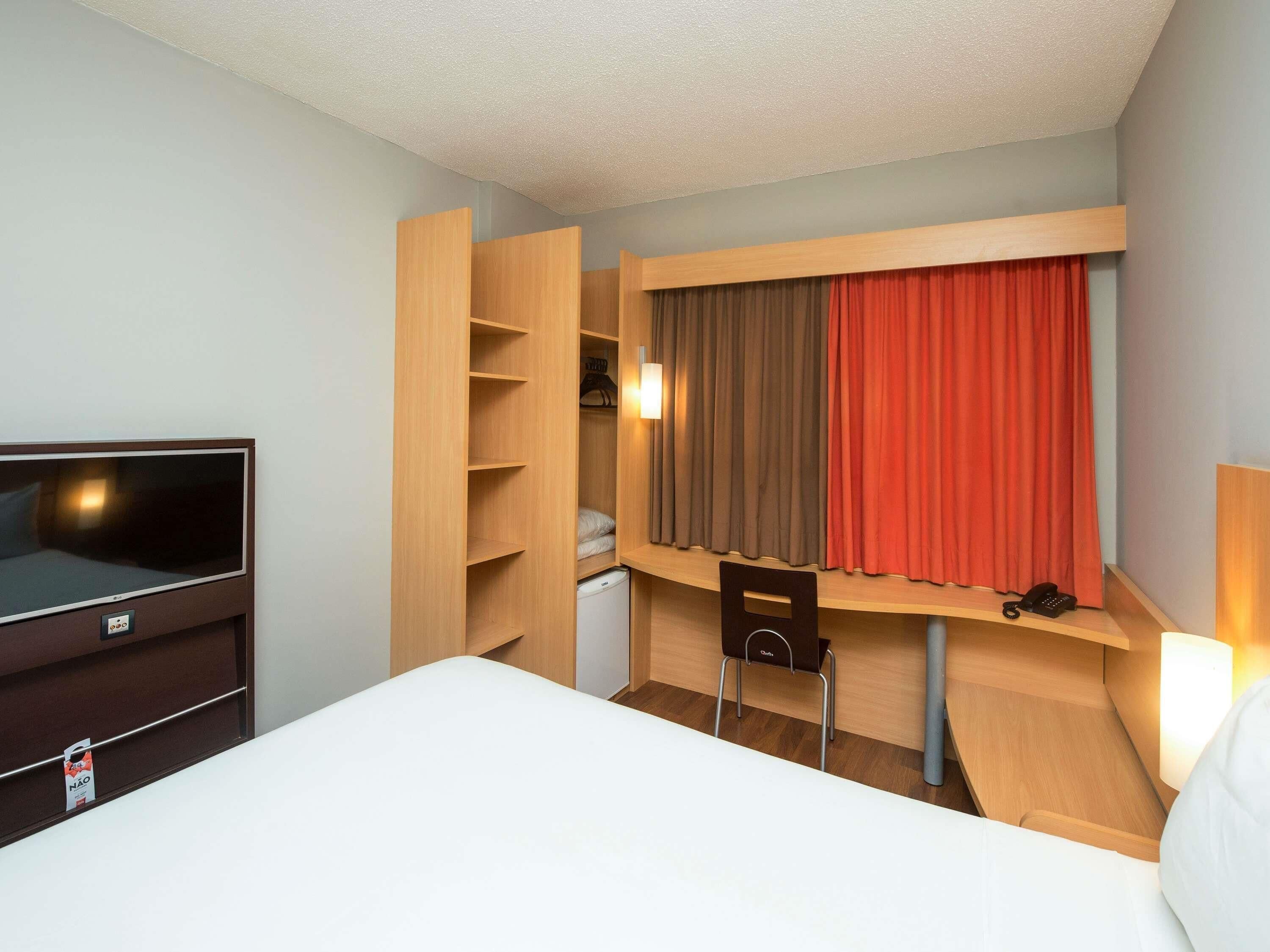 Une chambre d’hôtel comprenant un lit, une télévision, une armoire en bois, un bureau avec une chaise et une fenêtre avec des rideaux.