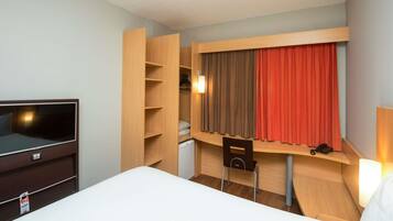 A hotel room with a bed, a TV, a wooden wardrobe, a desk with a chair, and a window with curtains.