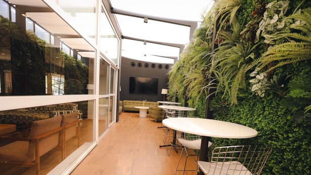 Un espace intérieur moderne doté d’un mur de verre, d’un plancher en bois et d’un mur végétal. On y trouve des coins salon avec des tables et des chaises.