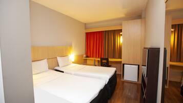 A hotel room with two beds, a desk, a chair, a small refrigerator, and a wardrobe.