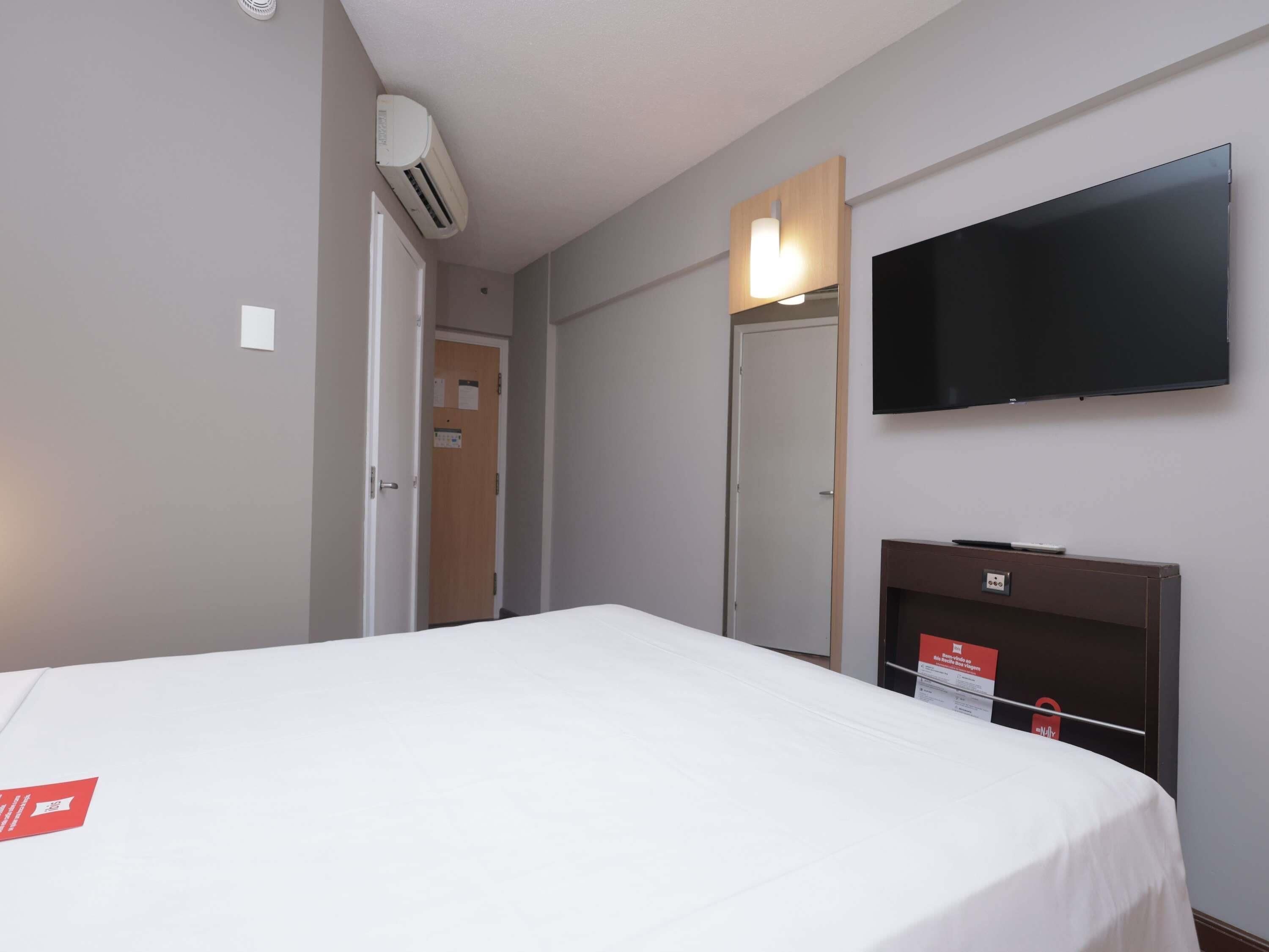 A hotel room with a bed, a TV mounted on the wall, an air conditioning unit, and a door leading to another room.