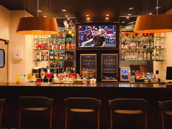 Uma área de bar com uma tela de televisão, prateleiras com garrafas de bebidas e banquetas.