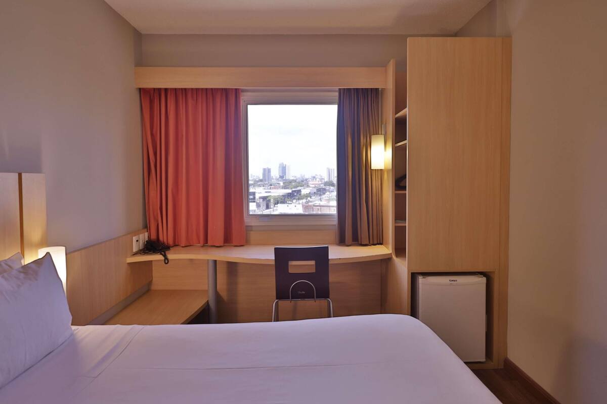 Une chambre d’hôtel équipée d’un lit, d’un bureau, d’une chaise, d’un petit réfrigérateur et d’une fenêtre avec des rideaux.