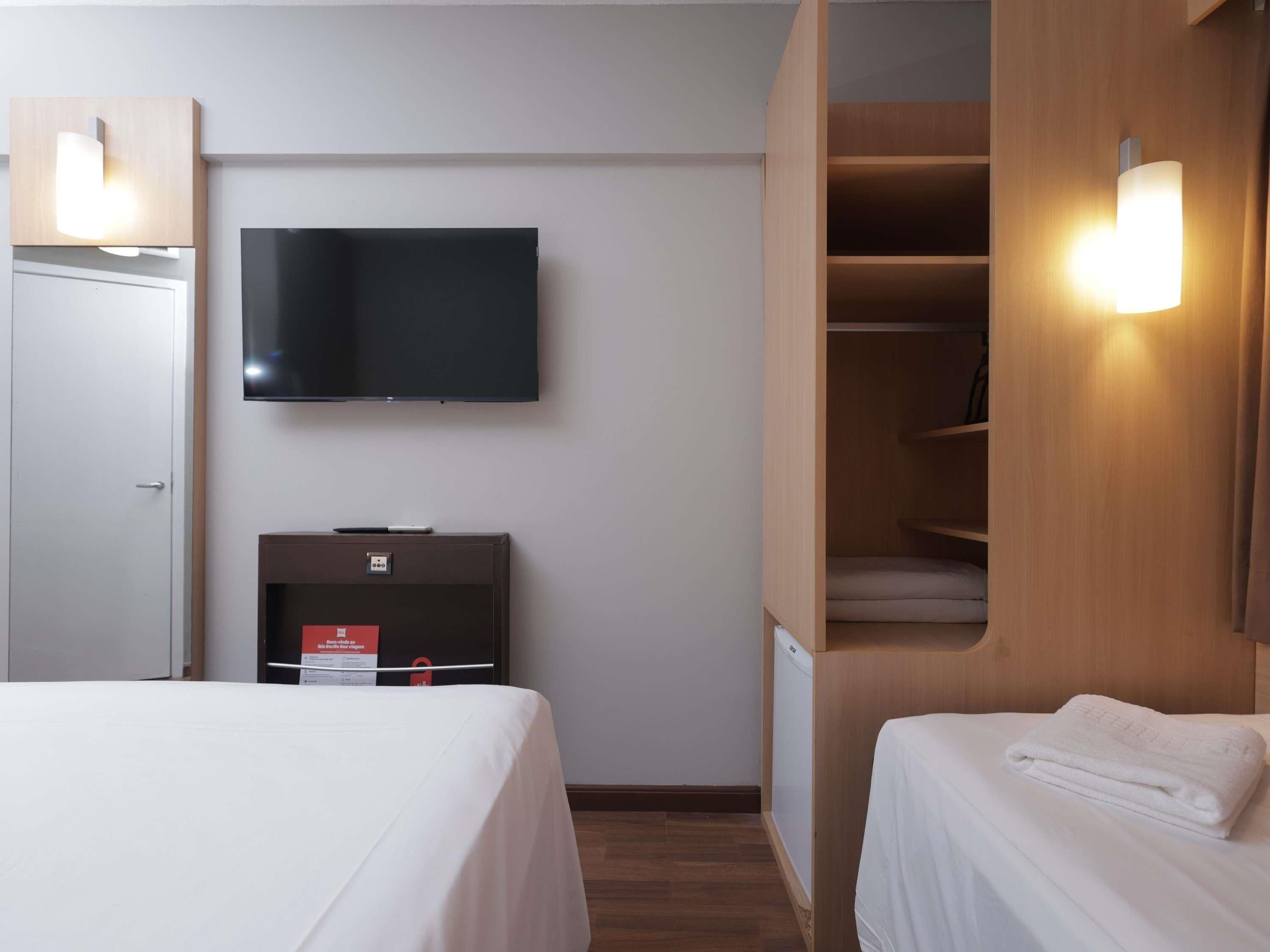 Hotellihuone, jossa on seinälle asennettu taulutelevisio, pieni televisio jalustalla, peilillä varustettu vaatekaappi ja kaksi sänkyä valkoisilla vuodevaatteilla.