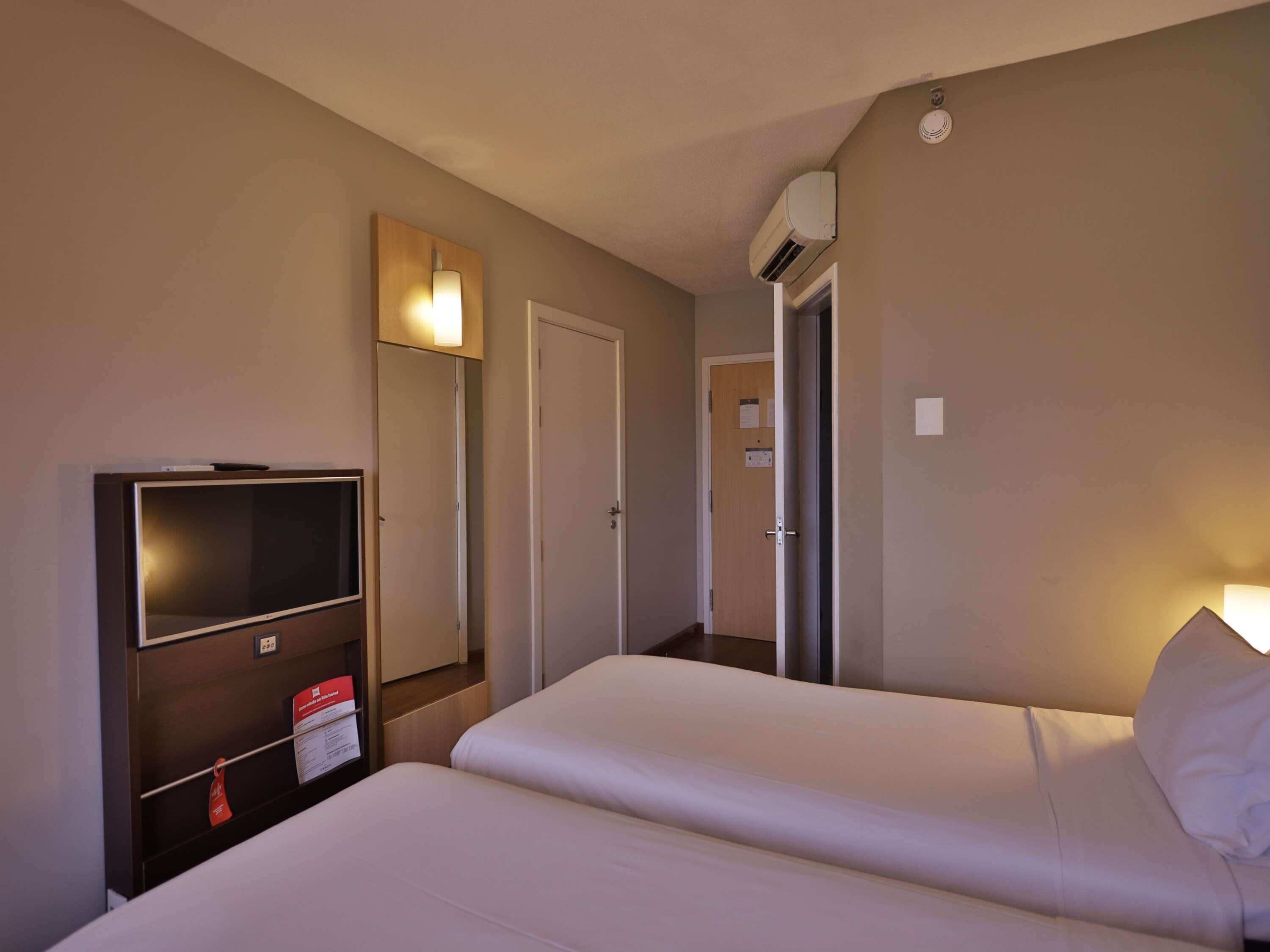 A hotel room with two beds, a TV, a mirror, and an air conditioning unit.