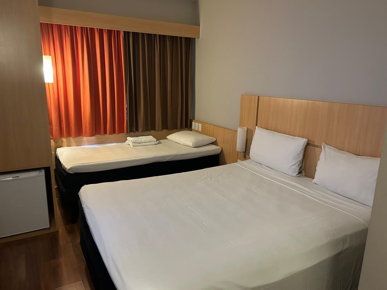 Habitación de hotel con dos camas, un refrigerador pequeño y una ventana con cortinas.