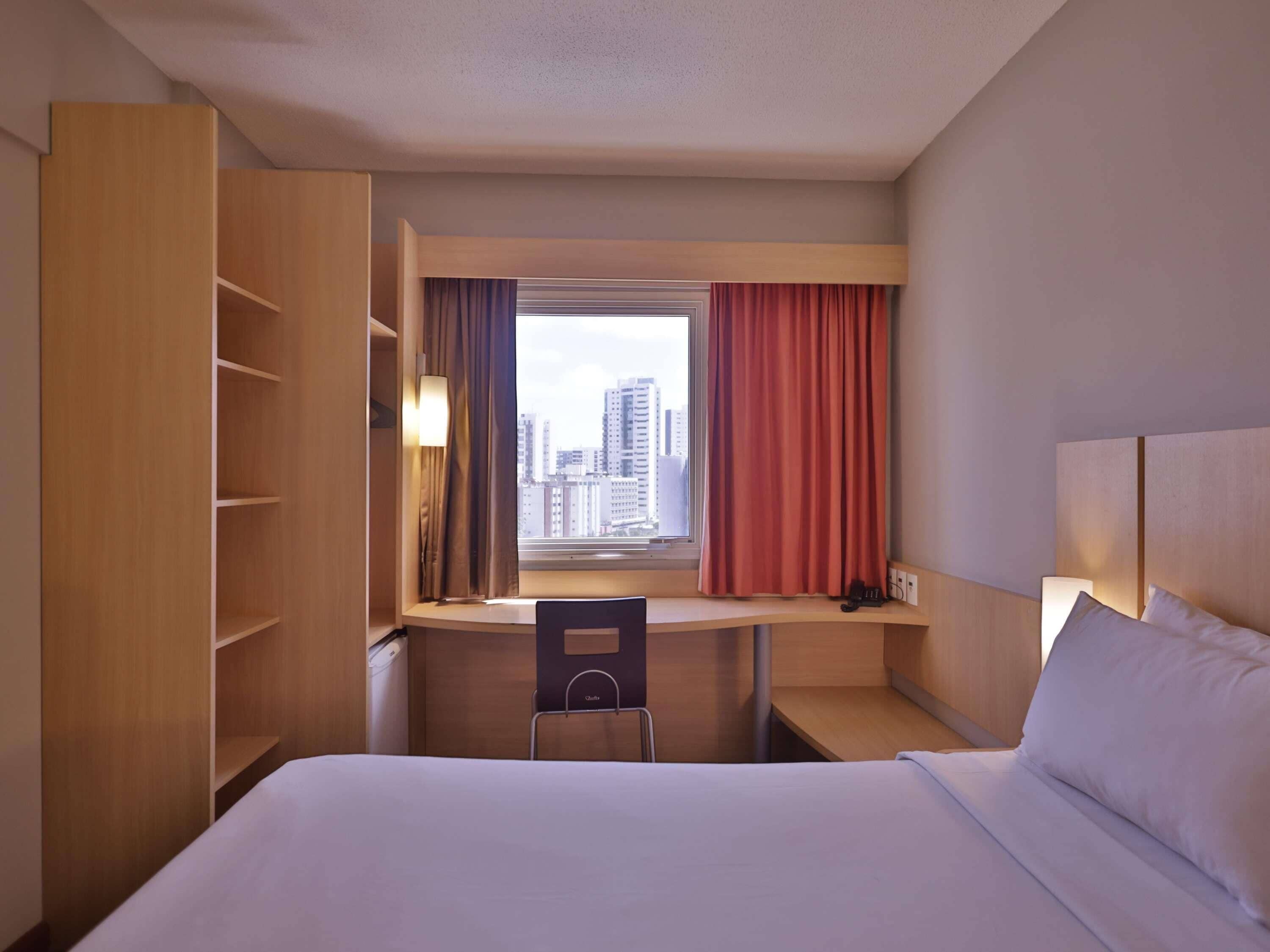 Habitación de hotel con cama, escritorio, silla, armario, ventana con cortinas y vista a la ciudad.