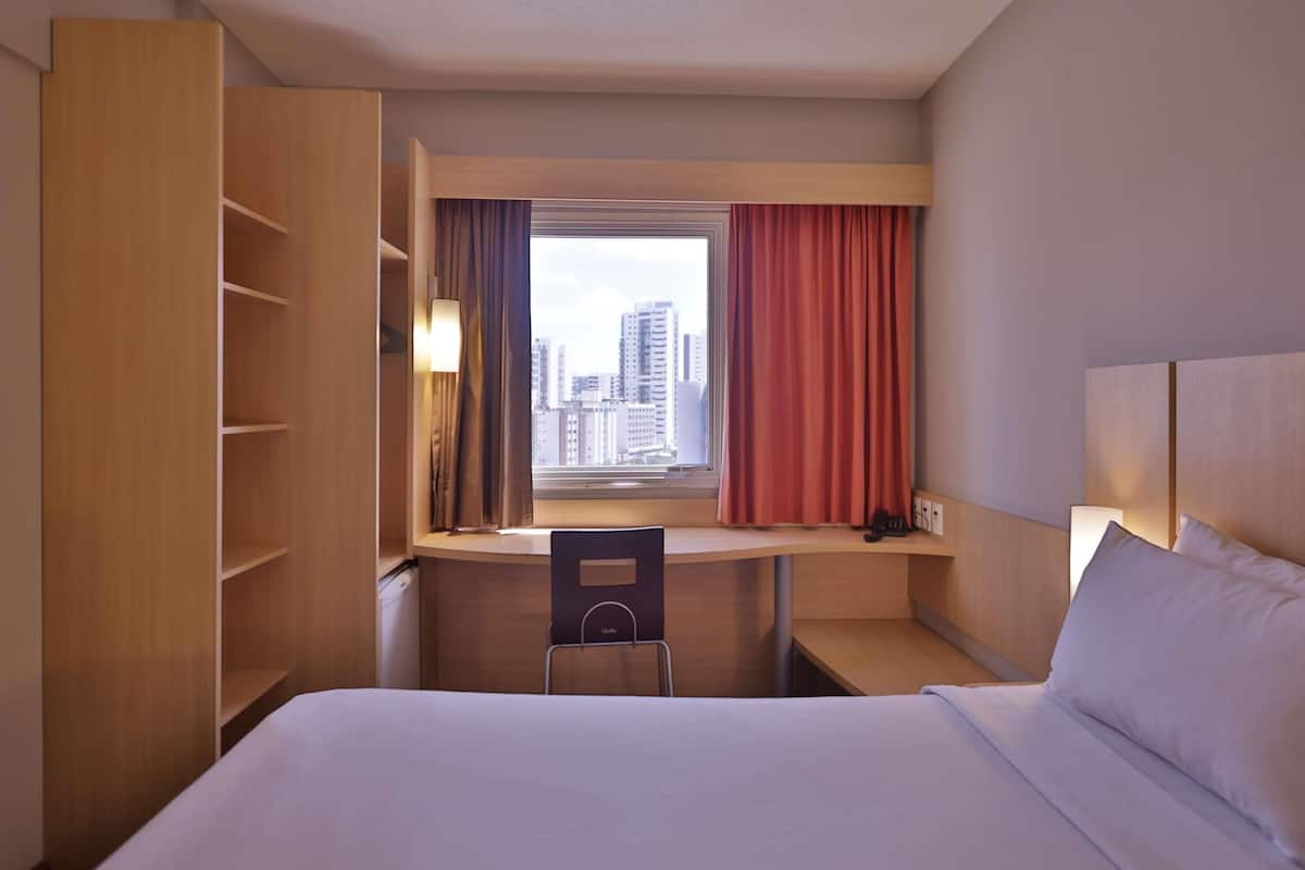 Habitación de hotel con cama, escritorio, silla, armario, ventana con cortinas y vista a la ciudad.