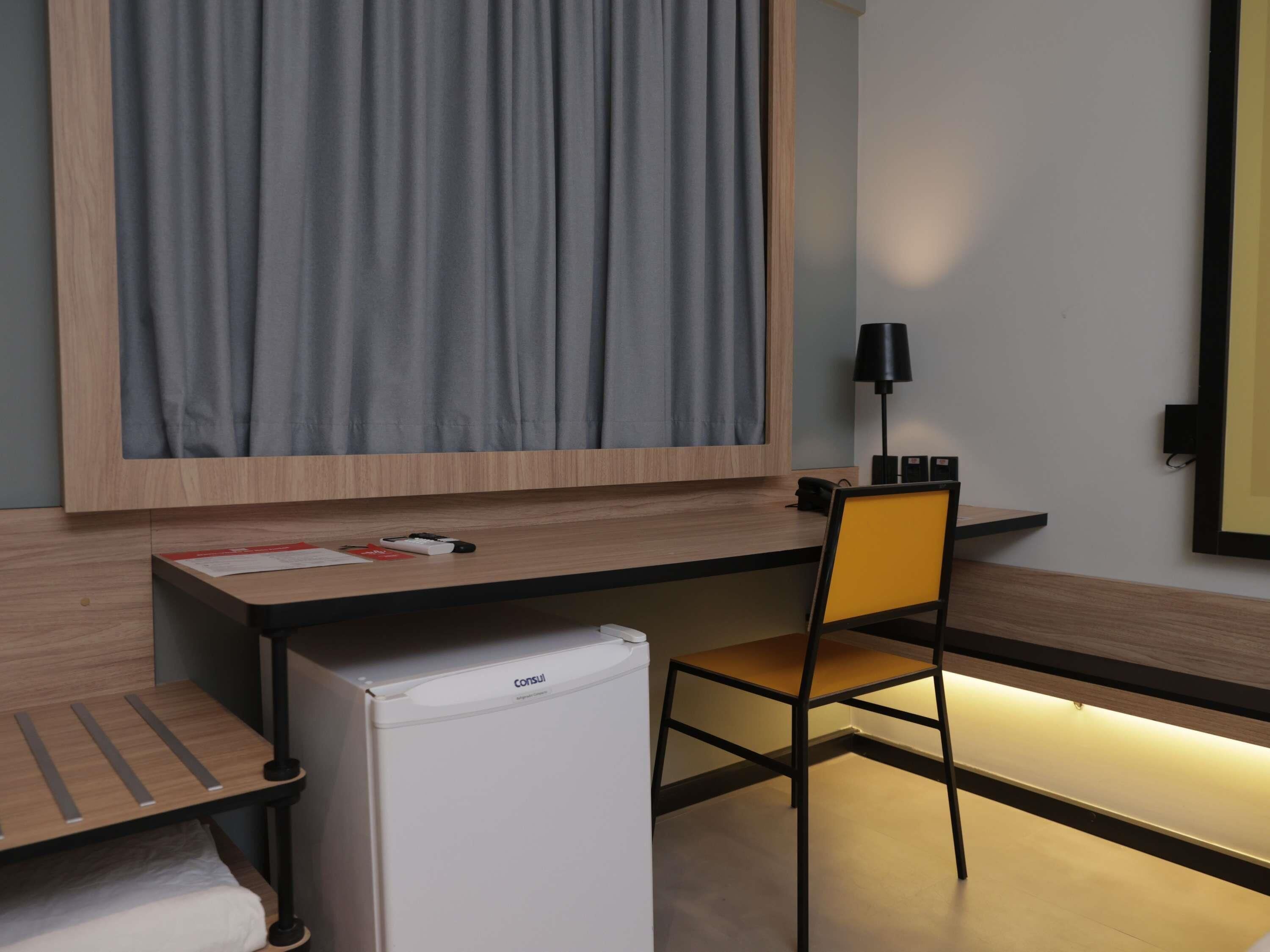 A modern hotel room with a wooden desk, a chair, a small refrigerator, and a large window with curtains.