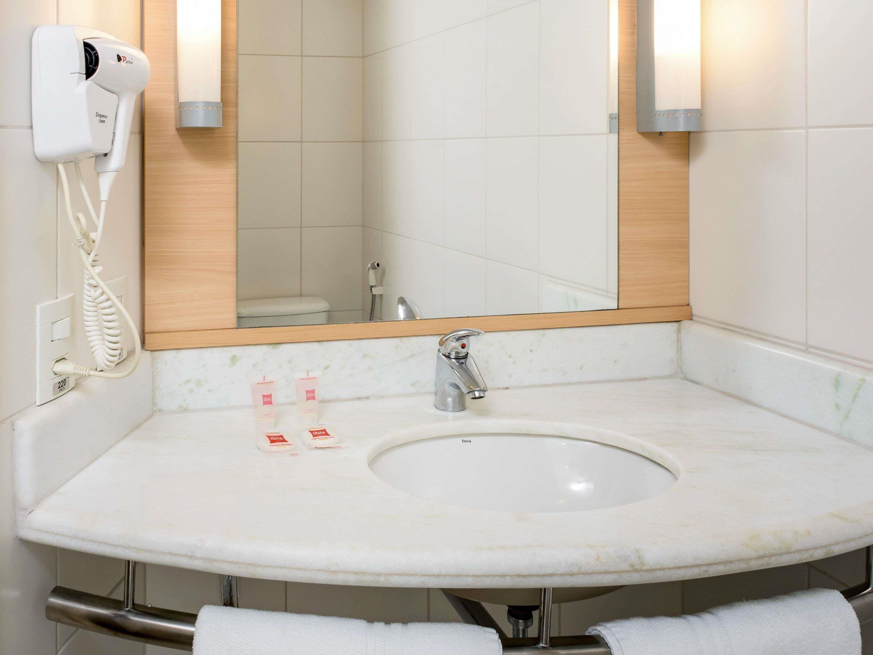 Un baño con lavamanos, espejo, toallero y secador de pelo fijado en la pared.